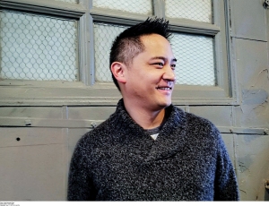 Photo of Mike Chen, an Asian man wearing a black sweater standing in front of a gray wall with block windows