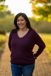 Author Photo: Lisa Sherman, woman wearing jeans and sweater standing in front of blurred outdoor scene
