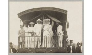 Sepia-toned photograph of old train, with several people in light-colored, early 20th-century clothing standing on the rear platform of the train.