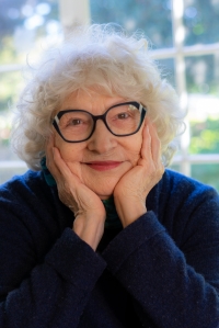 Author Norma Watkins photograph: smiling older white woman with short silvery-platinum colored hair, wearing black eyeglasses and a dark top with both hands cupping her face.