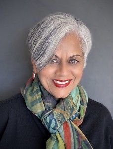 Alka Joshi author photograph: headshot of a smiling East Asian Indian women with short hair gray, wearing a black top and a multicolored neck scarf