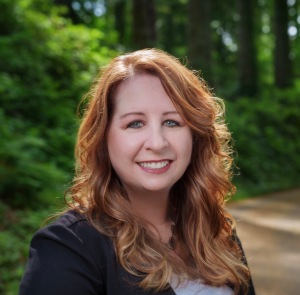 Author JL Lycette Photo: headshot of a red-haired smiling female standing in front of a wooded background