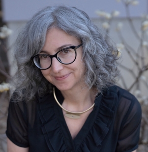 Photo of Marsha Gordon, picture of a white female wearing black eyeglasses, a gold statement necklace, short-sleeve black top, and graying, wavy, shoulder-length hair.