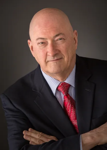 Photo of Kim Megahee, older, bald, white male with arms crossed, wearing a dark suit, light colored shirt, and red tie.