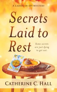 SECRETS LAID TO REST by Catherine C Hall book cover featuring a golden background, a small round table with a pumpkin pie centered on top and a "card" propped up against the pie