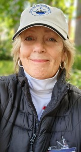 Author D MacNeill Parker photograph: older smiling White female with short blonde hair, wearing a ball cap, white turtleneck and a black quilted jacket/vest