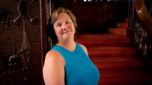 Author P.A. DePaul photo: white female with short brown hair wearing a turquoise sleeveless top standing with her back to a wall and red-carpeted steps in the background