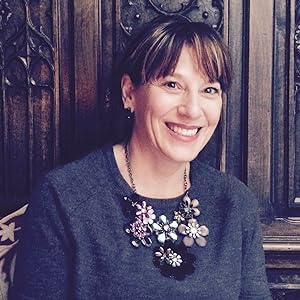 Photo of Andrea Rubinowitz, a smiling white female with highlighted brown hair, wearing a blue shirt and a decorative oversized floral necklace in front of a decorated wooden wall